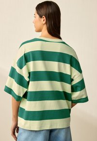 Tricou cu dungi verzi și verde deschis, croială lejeră, mâneci scurte, decolteu rotund, material moale din bumbac, purtat cu pantaloni scurți din denim deschis la culoare.