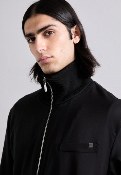 Black zip-up jacket with a high collar, flat pocket detail, textured fabric, and silver hardware for the zipper.
