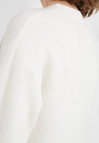 Witte, zachte gebreide trui met een ronde halslijn en verlaagde schouders. De stof heeft een subtiele textuur met een lichte glans.