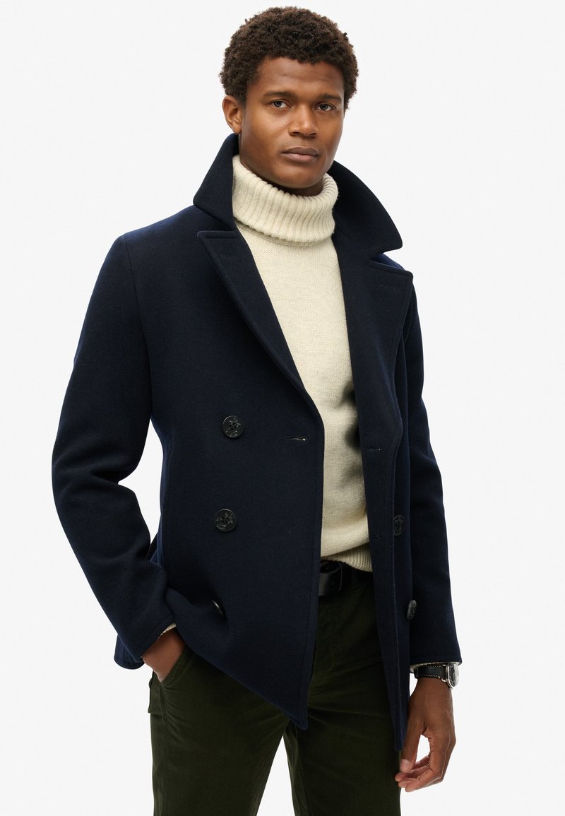 Marineblå uld peacoat med dobbeltbrystede knapper og brede revers, båret over en cremefarvet turtleneck, parret med mørkegrønne bukser.