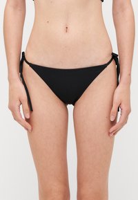 Fekete bikini alsó, amely oldalt megkötős záródással rendelkezik, alacsony derekú fazon, sima textúrával és minimalista dizájnnal. Egybefüggő stílus, minták nélkül.