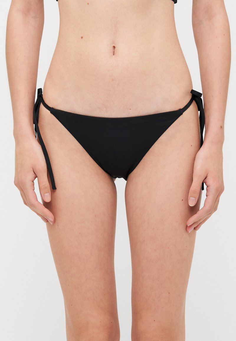 Fekete bikini alsó, amely oldalt megkötős záródással rendelkezik, alacsony derekú fazon, sima textúrával és minimalista dizájnnal. Egybefüggő stílus, minták nélkül.