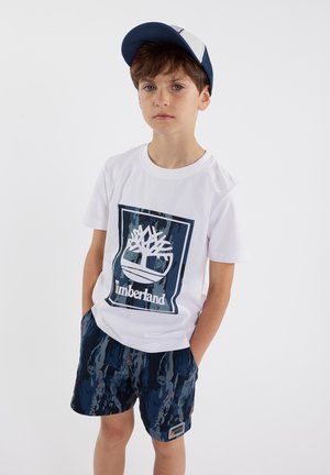 T-shirt blanche à manches courtes avec logo Timberland bleu, associée à un short à motifs bleu marine foncé et une casquette bleu marine avec panneau en maille blanche.
