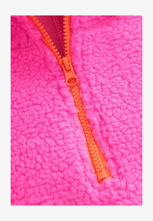 Fleece pulover v svetlo rožnati barvi z teksturirano površino, ki ima oranžno zadrgo in šivane podrobnosti vzdolž šivov.