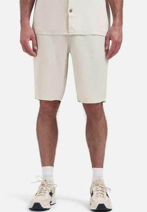 Man draagt beige shorts, witte sokken en beige sneakers en staat tegen een effen lichte achtergrond.