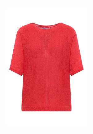Femme debout portant un pull rouge à manches courtes, un jean court bleu clair et des chaussures marron sans lacets sur un fond beige.