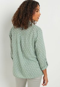 Blusa a maniche lunghe verde chiaro con pochette rettangolari bianche, dalla consistenza morbida e dal taglio rilassato, con plissettatura sul retro.