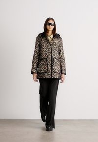 Pletená puffer bunda v leopardím vzoru s kapucí, dvěma předními kapsami a černými detaily; nošena přes béžovou košili a černé proužkované kalhoty.