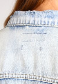Light blue denim jacket back view, featuring a standard collar, stitching details, and a small rectangular patch near the neckline.