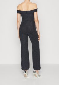 Off-the-shoulder haalari tummansinisena, jossa on metallinen kiilto, vartaloa myötäilevä siluetti, leveät lahkeet ja vetoketj closure selässä. Yhdistetty hopeisiin korkokenkiin.