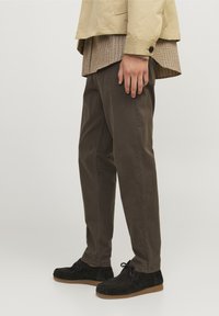 Personne portant un pantalon marron, des chaussures en daim noires à lacets, une veste beige et une chemise à carreaux, debout devant un fond uni.