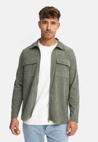 Mand iført en lys grøn kordjakke med lynlås over en hvid T-shirt og lyseblå jeans, stående foran en ensfarvet baggrund.
