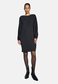 Zwarte jurk met lange, pofmouwen en een ronde hals, gemaakt van gladde stof, knielengte met een rechte snit. Draagt zwarte enkellaarsjes.