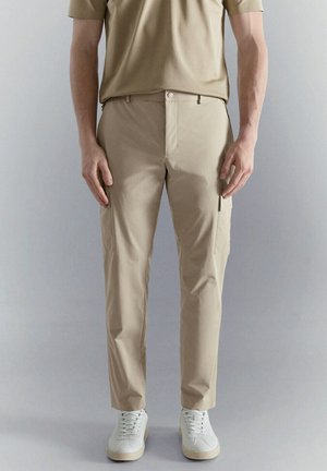 Hombre con pantalones casuales beige y camisa a juego, de pie con los brazos relajados a los lados frente a un fondo gris liso.