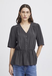 Schwarzes Peplum-Top mit kurzen Ärmeln, V-Ausschnitt, Knöpfen an der Vorderseite und geraffter Taille. Glatter Stoff mit dezentem Glanz. Kombiniert mit Jeans.