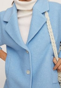 Abrigo de lana azul claro con cuello de muesca, bolsillos frontales y botones transparentes, superpuesto a un cuello alto blanco con una correa de crema texturizada.