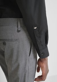 Crna košulja dugih rukava s manžetom na gumbima uparena je s sivim kariranim hlačama uskog kroja koje imaju suptilnu teksturu.