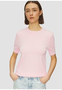 Lichtroze t-shirt met korte mouwen en een ronde hals, gemaakt van een gladde, zachte stof. Licht getailleerd, getoond met handen in de zakken van blauwe jeans.