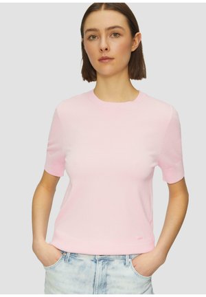Lichtroze t-shirt met korte mouwen en een ronde hals, gemaakt van een gladde, zachte stof. Licht getailleerd, getoond met handen in de zakken van blauwe jeans.