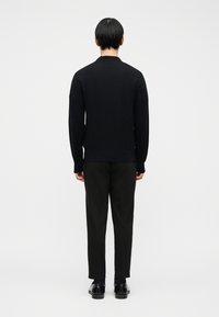Άνδρας που στέκεται έχοντας γυρισμένη την πλάτη, φοράει μαύρο πουλόβερ με μακριά μανίκια, μαύρο παντελόνι, μαύρες κάλτσες και μαύρα παπούτσια επίσημου τύπου, μπροστά σε λευκό μονόχρωμο φόντο.