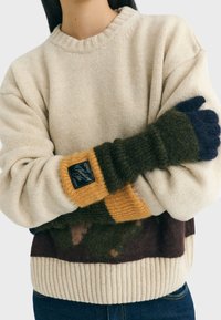 Sweter dziergany w kolorze kremowym, z prążkowanym kołnierzem i dołem. Kolorowe mankiety w odcieniach oliwkowym, brązowym i musztardowym, z naszywką na rękawie.