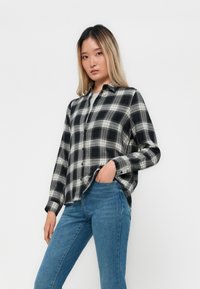 Svart og hvitt rutete flanellskjorte med knapper foran, lange ermer og avslappet passform, kombinert med blå denimjeans.