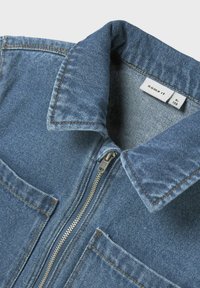 Denim jakna u svijetloplavoj boji, s oštrim ovratnikom, prednjim džepovima i zatvaranjem na zatvarač. Istaknuti detalji šivanja su prisutni.