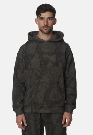 Man met kort donker haar, gekleed in een donker hoodie met camouflagepatroon en bijpassende broek, staand tegen een effen lichte achtergrond.