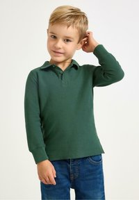 Zelená polokošile s dlouhým rukávem a límečkem, s dvoubotkovou légou a žebrovanými manžetami, kombinovaná s modrými džíny. Hladká textura a přiléhavý střih.