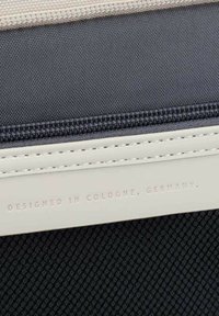 Zapínací taška z tmavé síťované látky s krémovým lemem. Obsahuje embosovaný text: „DESIGNOVÁNO V KOLE KAMENICE, NĚMECKO.“ Odolný, strukturovaný povrch.