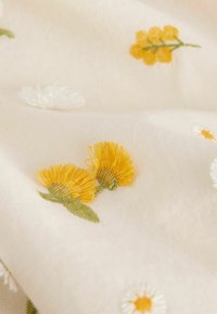 Tejido de algodón con intrincados bordados florales en amarillo y blanco. Presenta una textura suave y un fondo color crema claro.