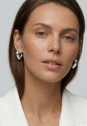 Kvinde med lys hud og glat brunt hår bærer skinnende, sølvfarvede, tykke øreringe formet som hjerter og en hvid blazer mod en lys baggrund.