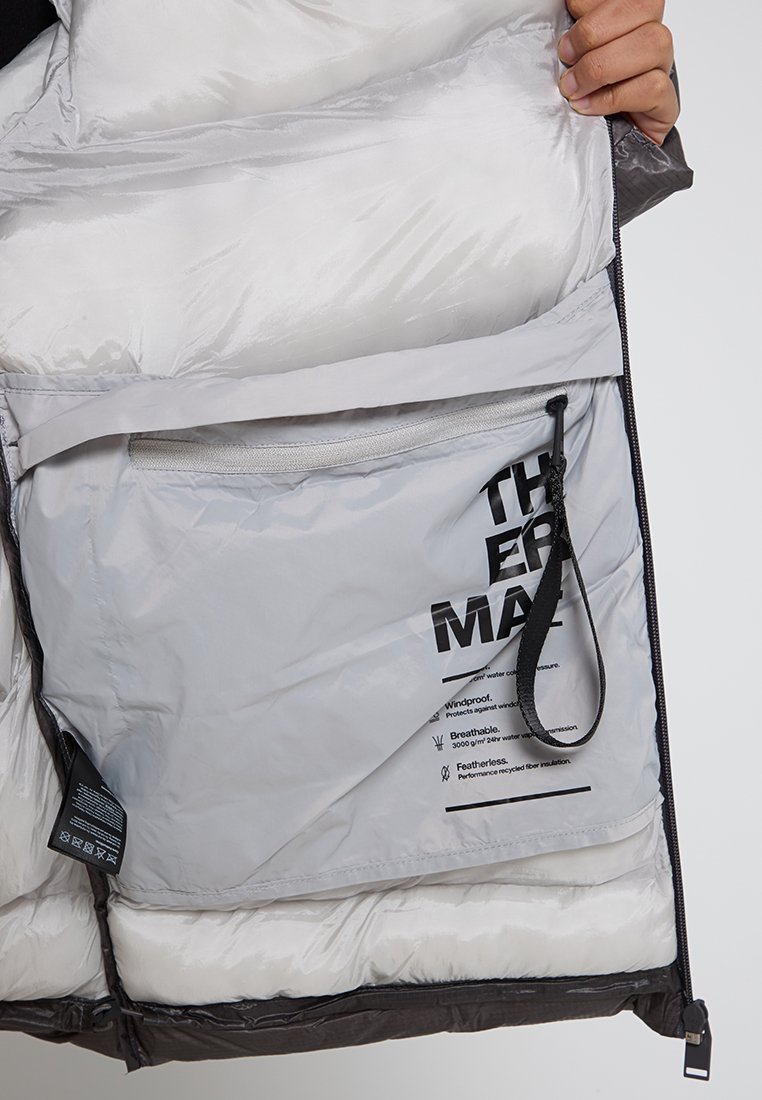 Giacca isolante con un guscio esterno nero e un interno grigio chiaro. Presenta una tasca con zip e etichette che dettagliamo le proprietà del tessuto.