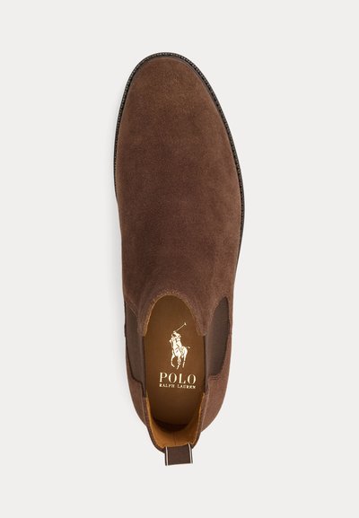 Barna szarvasbőr bebújós cipő elasztikus oldalsó panelekkel és húzófüllel. Arany logóval díszített világosbarna talpbetét és strukturált forma jellemzi.