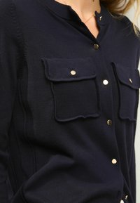 Navy knit cardigan featuring two front pockets with flap and button closures. Gold buttons accent the front. Soft texture, fitted design.