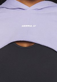 Gros plan d'une personne portant un sweat à capuche court lilas avec le logo "NEBBIA" sur un haut noir, montrant un petit détail d'ouverture poitrine.