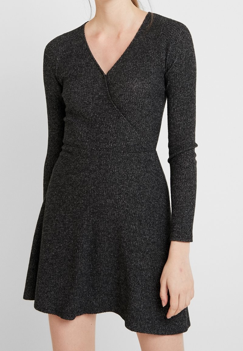 Svart ribbad klänning med V-hals och långa ärmar, åtsittande midja och utsvängd kjol, med mjuk textur och minimalistisk design.