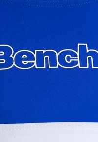 Modra športna crop majica z izstopajočim belim logotipom "Bench". Gladka tkaninska tekstura z kontrastnim belim robom na dnu.