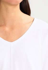 Vit V-ringad topp med en mjuk textur. Har en lätt axeldetalj, rena linjer och en subtil söm fram.