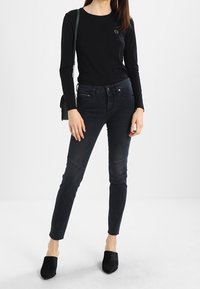 Černý dlouhý rukáv s malým logem, v kombinaci s tmavými džíny s úzkým střihem a detaily na kolenech, doplněné černými muly.
