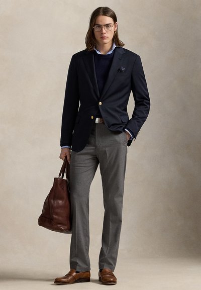 Mand med langt hår og briller iført navy blazer, grå bukser, brune sko, der holder en stor brun lædertaske mod en neutral baggrund.
