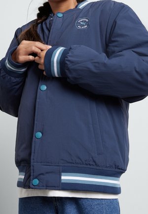 Marineblaue Bomberjacke mit rundem Aufnäher, gerippten Bündchen mit blau-weißen Streifen, Druckknöpfen und glattem, leichtem Stoff.
