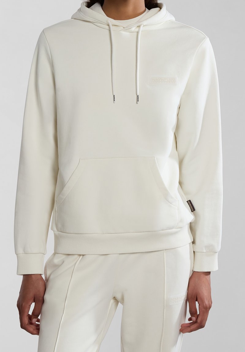 Personne portant un sweat à capuche crème avec poche frontale et cordons, assorti à un pantalon coordonné sur un fond neutre.