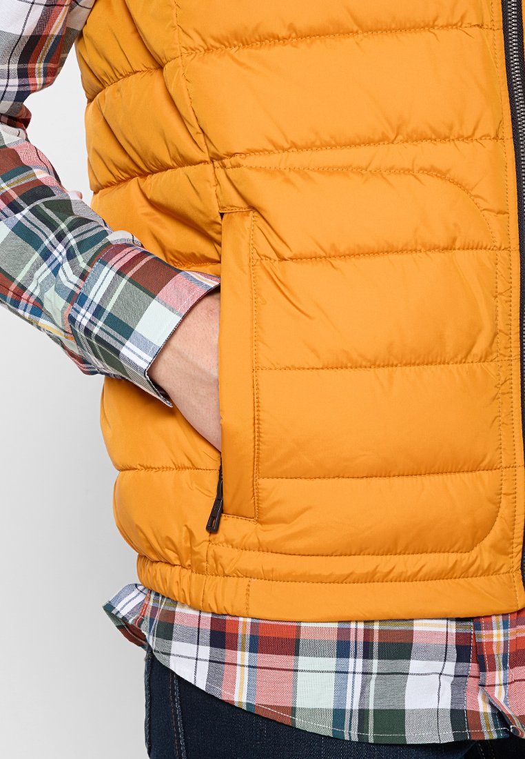 Un gilet rembourré jaune avec des coutures horizontales et une poche latérale, porté sur une chemise à carreaux multicolore comportant des tons rouges, verts et bleus.