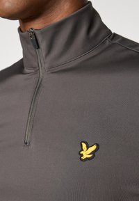 Mørkegrå halv-zip pullover lavet af strækstof, med et broderet gult fuglelogo på venstre bryst. Glat tekstur.