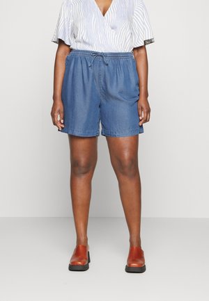 Person trägt blaue Denim-Shorts mit elastischem Bund und Kordelzug, ein weiß-blau gestreiftes Kurzarmhemd und braune Slipper.