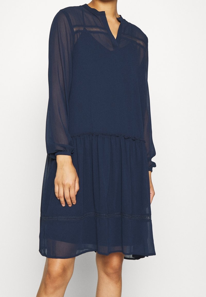 Femme portant une robe bleu marine à manches longues, longueur genou, avec des manches transparentes et un détail froncé à la taille.