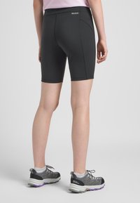 Pantaloni corti sportivi neri con un design aderente, texture liscia e vita alta. Modella che indossa scarpe da trekking grigie con dettagli viola.