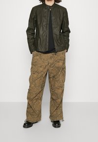 Mörkgrön läderjacka med ståkrage, kombinerad med löst sittande bruna cargobyxor med en strukturerad, mönstrad design. Svarta stövlar nedanför.