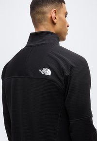 Άνδρας που φοράει μαύρο μπουφάν North Face, εμφανιζόμενος από πίσω και πλάγια, με φόντο ένα απλό ανοιχτό γκρι.